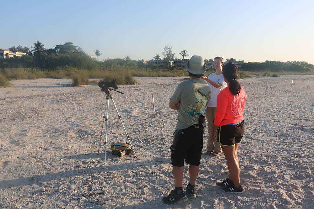 Three people stands conversing on a beach next to a monocular on a tripod.